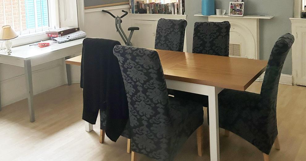 A sizeable dining area space, centrally to the room is a large six person wooden dining table with fleur de lis covered grey and black chairs. Under the window sits a white side table. The room is neutral and uses the natural light from the window and wooden flooring to create an airy space.