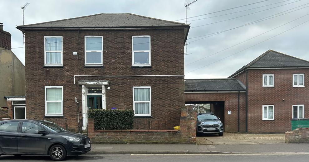 A detached, two storey brown brick built property, with a small brick built wall around the perimeter of the main building. Joined onto the building is a covered opening housing a parking space and access to the back of the property.