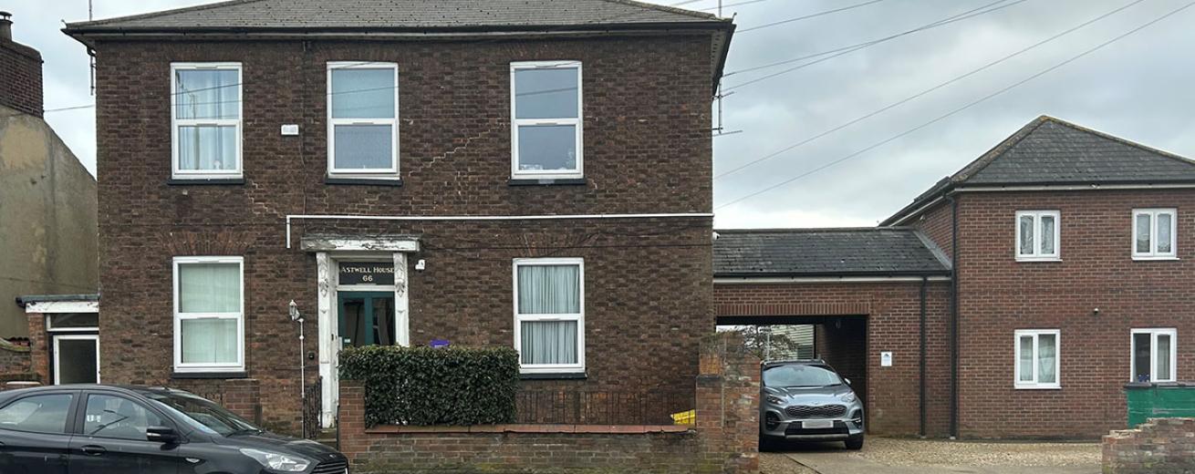 A detached, two storey brown brick built property, with a small brick built wall around the perimeter of the main building. Joined onto the building is a covered opening housing a parking space and access to the back of the property.
