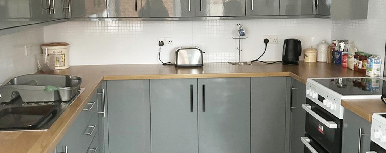 A modern high gloss grey kitchen cabinetry decorated kitchen, broken up with white walls, a natural wood work top. Chrome kitchen accessories and two white electric hobs and ovens.