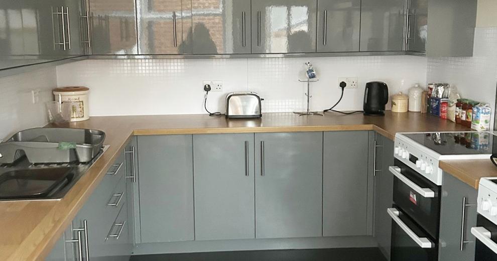 A modern high gloss grey kitchen cabinetry decorated kitchen, broken up with white walls, a natural wood work top. Chrome kitchen accessories and two white electric hobs and ovens.