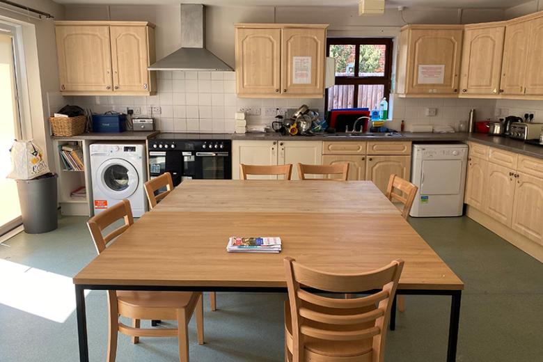 A large kitchen space with light wooden cabinetry forming a L shape of the room. A large range oven is fitted as well as washing machine and tumble dryer. In the centre of the room are two large wooden tables pushed together to create a large communal seating area.