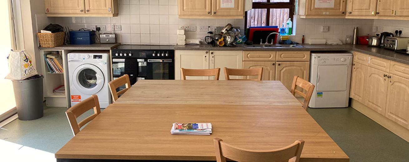 A large kitchen space with light wooden cabinetry forming a L shape of the room. A large range oven is fitted as well as washing machine and tumble dryer. In the centre of the room are two large wooden tables pushed together to create a large communal seating area.