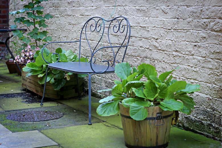 Metal framed two seater outdoor bench with a twisted metalwork design on the back panel. Potted planters sit adjacent to the bench in a row against the exterior wall of the property adding colour and natural textures to the paved garden.