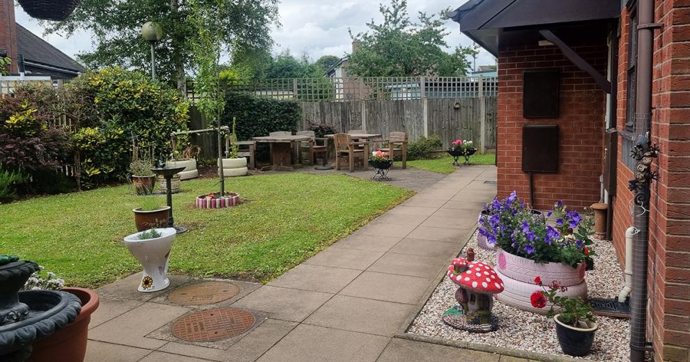 A compact and tidy garden area with a mixture of grassed and slabbed areas. The slabbed areas create a neat walkway to the multiple outdoor dining areas. With the grassed areas being well mainlined with a mature set of planted shrubs which line the fence. Decorative potted plants have been placed around the to inject pops of colour.