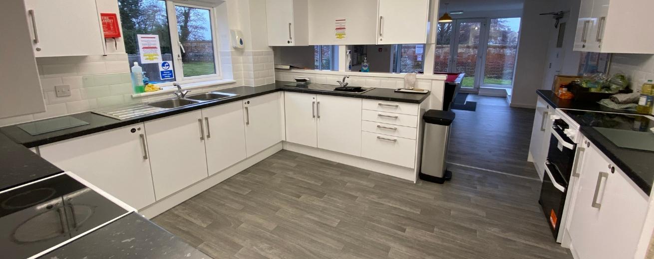 A well packed kitchen area, using a simple and clean white with dark worktops. The room has plenty of storage options as well as two white ovens and hobs, two stainless steel sinks and undercounter fridges.
