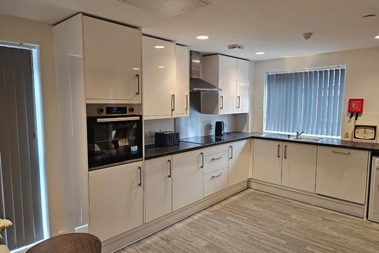 Modern communal kitchen with white gloss cabinets, black countertops, integrated oven, electric hob, and essential appliances like a toaster and kettle, with a large window and vertical blinds.
