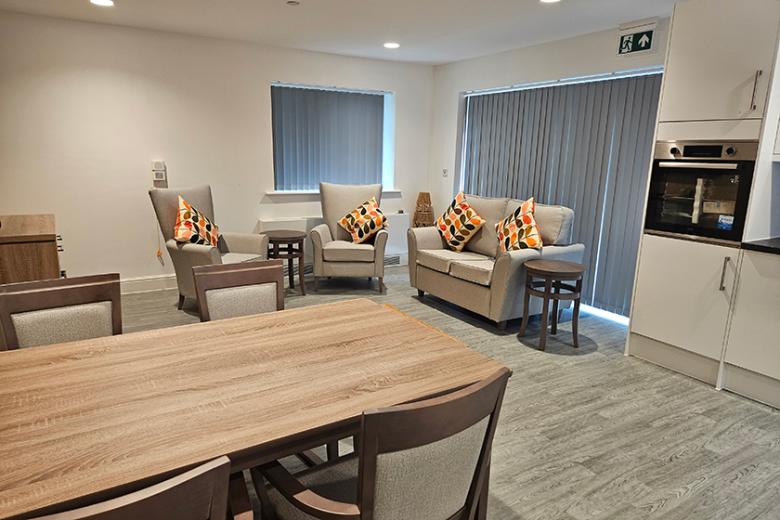 Communal living and dining area featuring a wooden dining table with six cushioned chairs, two armchairs, and a small sofa with patterned cushions, all set against white walls with grey blinds.