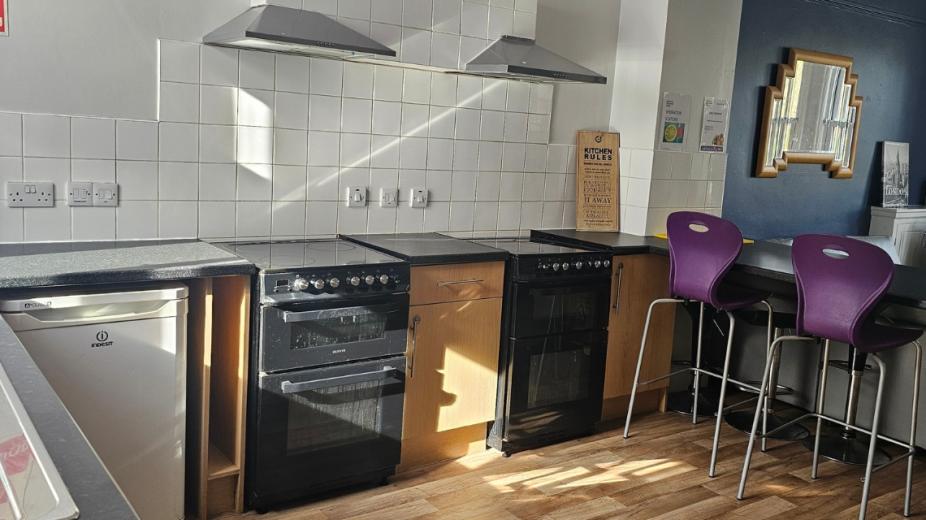 An earth toned minimalistic kitchen area with breakfast bar purple barstool seating, two black electric ovens and hobs an under counter fridge and an array of wood effect bottom cabinets.