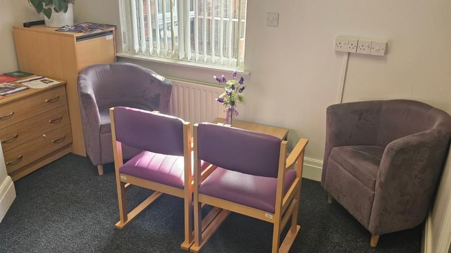 A compact brown carpeted seating area with two wood effect draws tucked into the corner and lining the wall of the room. A burgundy armchair faces two wooden chairs with lilac leather upholstery. A small wooden coffee table sits between them.