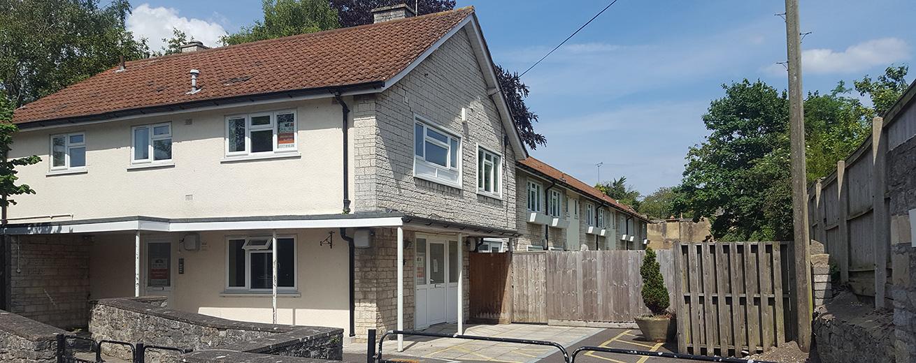 A two-story residential building with a light-colored exterior and a red-tiled roof is visible. In front of the building, there is a gated entrance leading to a parking area. The surrounding area features a stone wall and a wooden fence, with trees and greenery in the background. The sky is clear with a few clouds.