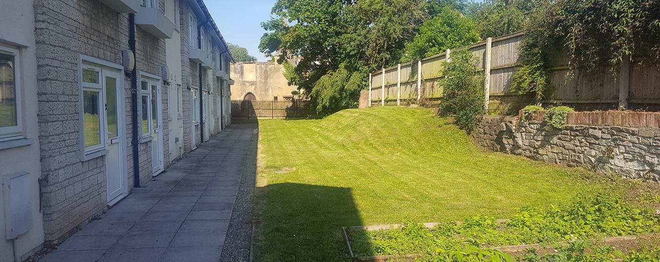 A well-maintained grassy area is bordered by a stone wall and a wooden fence. On the left, there are several residential units with white doors and windows, while the right side features a neatly trimmed lawn. The scene is bathed in sunlight, with trees and shrubs in the background, creating a peaceful outdoor environment.
