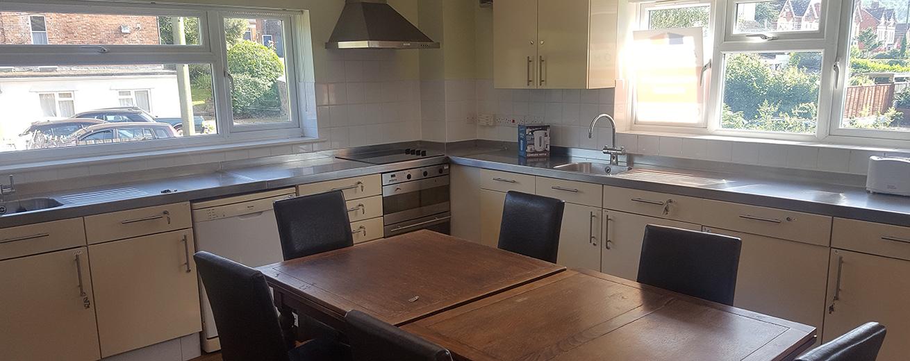 A spacious kitchen featuring light-colored cabinets and a stainless steel countertop. There is a large wooden dining table with black chairs in the center. The kitchen is well-lit by natural light coming through two windows, which offer a view of a garden outside. A stainless steel oven and a sink are visible along the counter, and a range hood is mounted above the stove.
