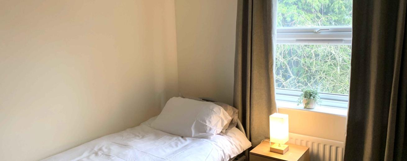 A cozy bedroom featuring a single bed with white bedding, positioned against a light-colored wall. Next to the bed is a wooden nightstand with a small lamp and a woven basket containing various items. A window with gray curtains allows natural light to enter, revealing greenery outside. The room has a simple and inviting atmosphere.
