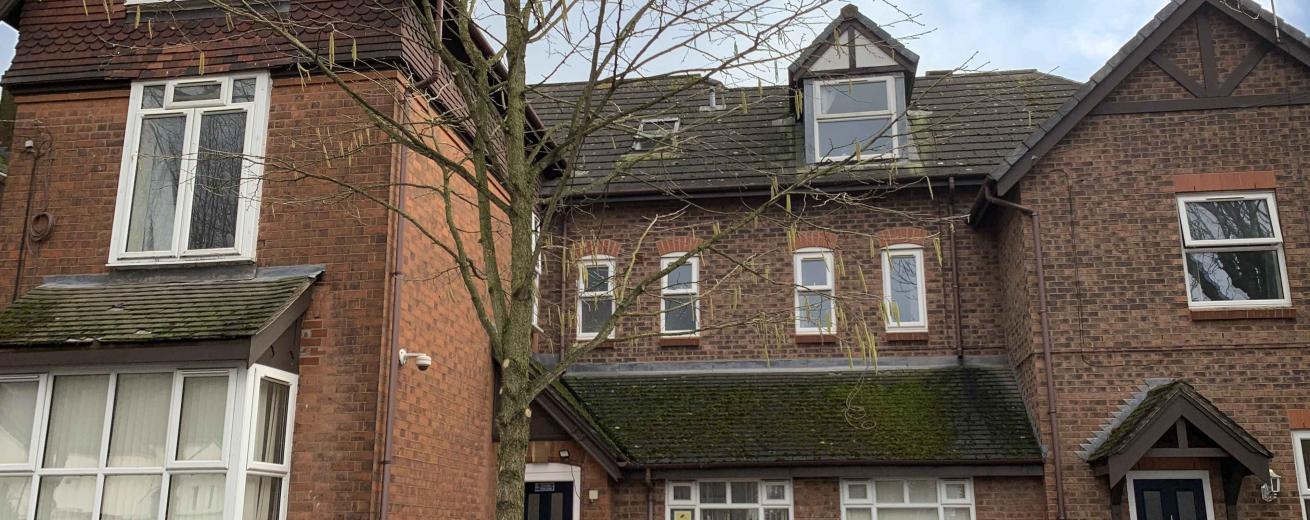 A brick residential building featuring multiple stories and a mix of window styles. The front yard has a small tree and two trash bins. A black car is parked in front of the building, and the sky above is partly cloudy.