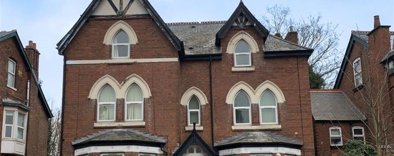 A large, three-story Victorian-style brick house with decorative gables and arched windows. The building features a symmetrical facade with a central entrance and two large bay windows on either side. The roof has a steep pitch, and there are trees in the background. A parked car is visible in front of the house, and the sky is partly cloudy.