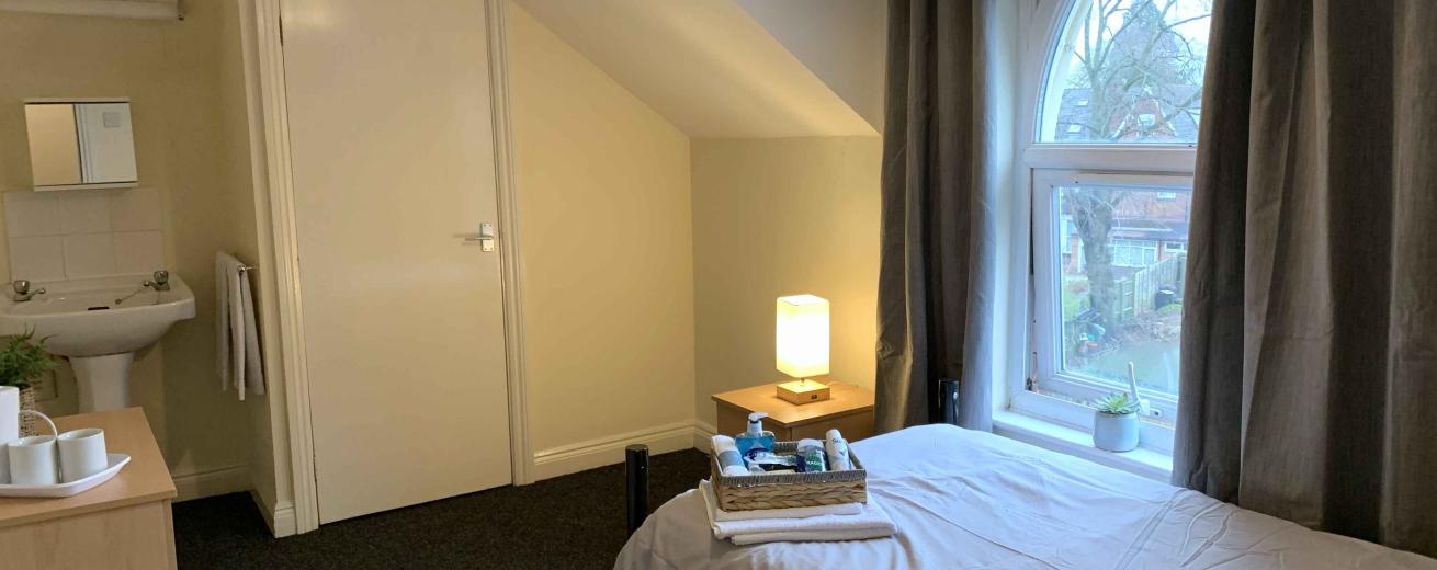 A cozy bedroom featuring a single bed with a neatly arranged white bedspread. Next to the bed is a small wooden bedside table with a lamp and a few toiletries. A window with an arched design allows natural light to enter, revealing a view of trees outside. The room has light-colored walls and dark carpet, with a door leading to a bathroom visible in the background. Gray curtains frame the window, adding a touch of warmth to the space.