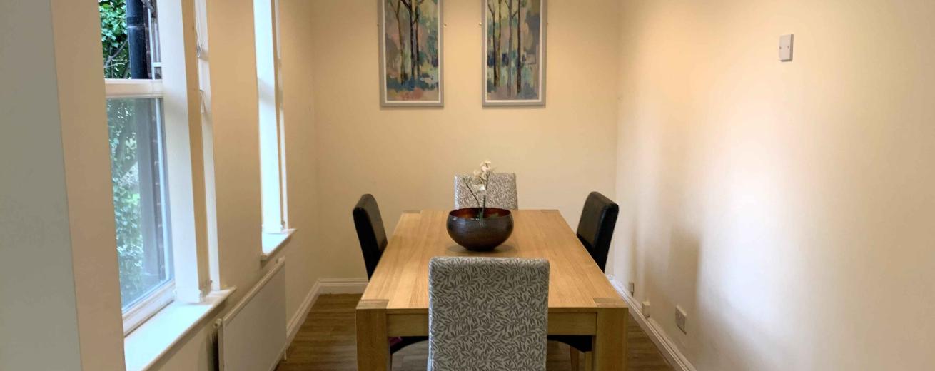 A bright and minimalistic dining area featuring a wooden table with a decorative bowl in the center. Surrounding the table are four chairs, two of which are upholstered in a light fabric. The walls are painted a soft cream color, and there are two framed artworks depicting trees hanging on the wall. Natural light streams in through two windows, enhancing the airy feel of the space.