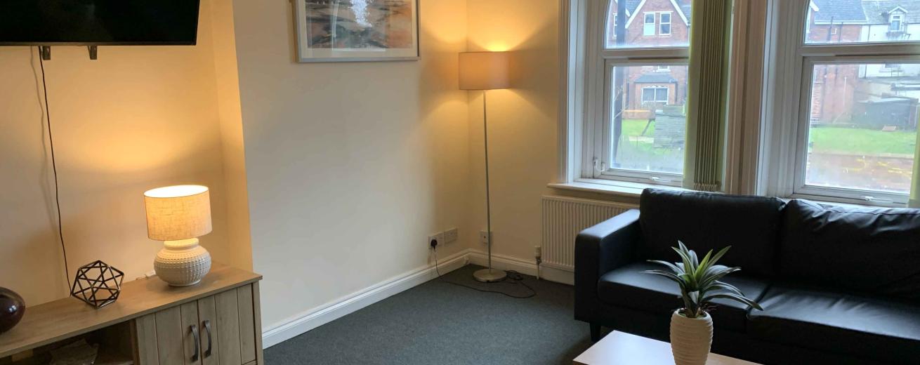A cozy living room featuring a dark sofa, a wooden coffee table, and a decorative plant. There is a television mounted on the wall and a lamp providing warm light. Large windows with arch-shaped tops allow natural light to enter, and a view of a rainy street can be seen outside. The walls are painted a soft yellow, and the carpet is dark gray.