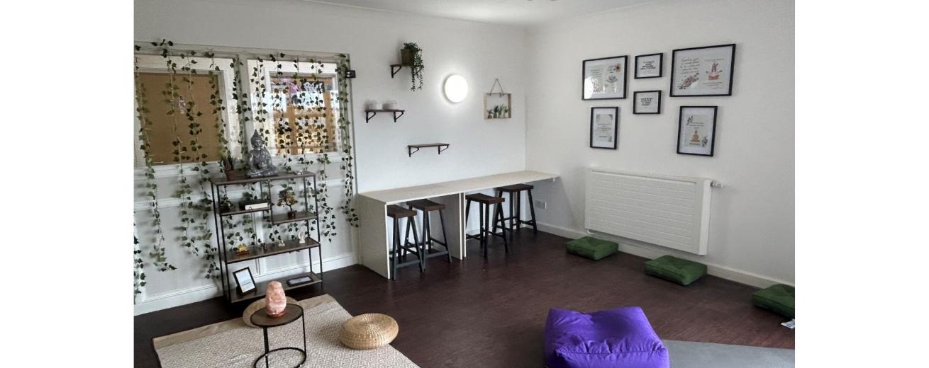 A serene and inviting room designed for relaxation and yoga. The space features a light-colored wall with decorative plants and framed artwork. On one side, there is a small table with two stools, and a shelf displaying a statue and plants. The floor is adorned with a large mat, two round cushions, and several green yoga blocks. Natural light enters through a window, enhancing the calm atmosphere.