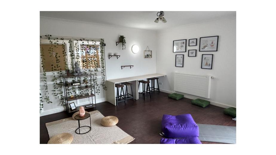 A serene and inviting room designed for relaxation and yoga. The space features a light-colored wall with decorative plants and framed artwork. On one side, there is a small table with two stools, and a shelf displaying a statue and plants. The floor is adorned with a large mat, two round cushions, and several green yoga blocks. Natural light enters through a window, enhancing the calm atmosphere.