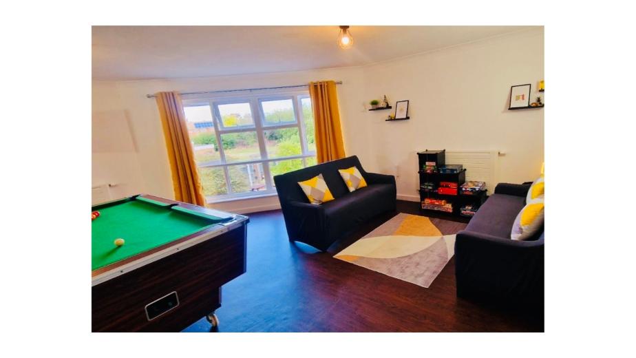 A cozy living space featuring a green felt pool table on the left, with a few colorful balls scattered on it. To the right, there are two dark sofas adorned with yellow and gray cushions, positioned around a light-colored area rug. A small bookshelf filled with games is against the wall, and large windows with yellow curtains allow natural light to fill the room. Decorative items are displayed on the walls and shelves, creating a welcoming atmosphere.