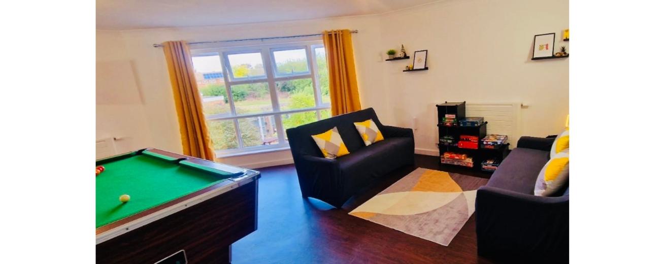 A cozy living space featuring a green felt pool table on the left, with a few colorful balls scattered on it. To the right, there are two dark sofas adorned with yellow and gray cushions, positioned around a light-colored area rug. A small bookshelf filled with games is against the wall, and large windows with yellow curtains allow natural light to fill the room. Decorative items are displayed on the walls and shelves, creating a welcoming atmosphere.