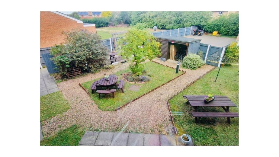 A view of a small garden from above, featuring a gravel pathway that winds through the greenery. There are wooden picnic tables on either side of the path, surrounded by grass and shrubs. A small tree is visible in the center, and a shed or storage unit is located at the back. The area is enclosed by wooden fencing, with a few trees and plants adding to the natural ambiance.