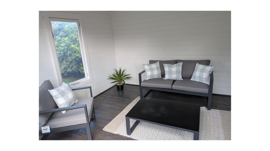A cozy indoor seating area featuring a gray sofa and armchair with decorative pillows, a black coffee table, and a light-colored rug. A small potted plant is placed beside the sofa, and a window offers a view of greenery outside. The walls are white, and the floor is dark wood.
