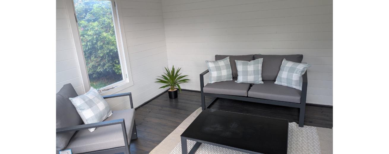 A cozy indoor seating area featuring a gray sofa and armchair with decorative pillows, a black coffee table, and a light-colored rug. A small potted plant is placed beside the sofa, and a window offers a view of greenery outside. The walls are white, and the floor is dark wood.