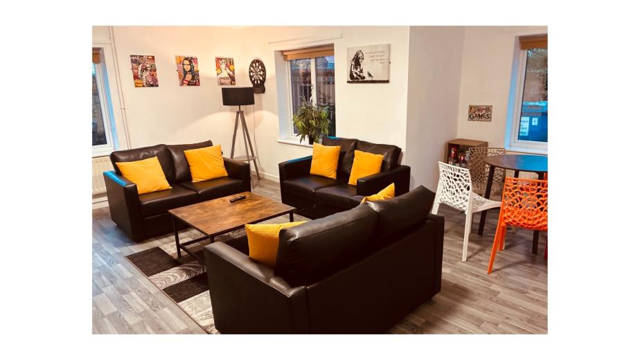 A cozy living room featuring two black sofas with bright yellow cushions, arranged around a wooden coffee table. A floor lamp stands in one corner, and a small dining table with colorful chairs is visible in the background. The walls are adorned with framed posters, and there are large windows allowing natural light to fill the space. A potted plant adds a touch of greenery to the room.