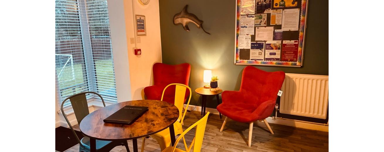 A cozy indoor seating area featuring two red armchairs and a round wooden table. The wall behind is painted dark green and adorned with a dolphin sculpture and a bulletin board filled with colorful notices. A small lamp sits on the table, providing warm light. Large windows allow natural light to fill the space, with a view of a grassy area outside. The floor is made of light wood, and there are metal chairs in various colors around the table.