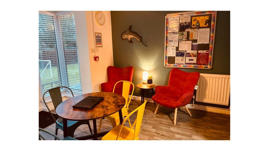 A cozy indoor seating area featuring two red armchairs and a round wooden table. The wall behind is painted dark green and adorned with a dolphin sculpture and a bulletin board filled with colorful notices. A small lamp sits on the table, providing warm light. Large windows allow natural light to fill the space, with a view of a grassy area outside. The floor is made of light wood, and there are metal chairs in various colors around the table.