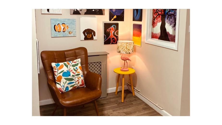 A cozy corner of a room featuring a brown leather armchair with a colorful floral pillow. Surrounding the chair is a gallery wall adorned with various artworks, including abstract pieces, animal portraits, and colorful landscapes. A small yellow side table holds a pink flamingo lamp, providing warm light to the space. The floor is wooden, and the overall atmosphere is inviting and artistic.