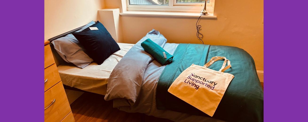 A cozy bedroom featuring a single bed with a blue and gray bedding set. A green towel is neatly placed on the bed, alongside a beige tote bag that reads "sanctuary supported living." A wooden dresser is visible on the left, and a window with natural light illuminates the room.