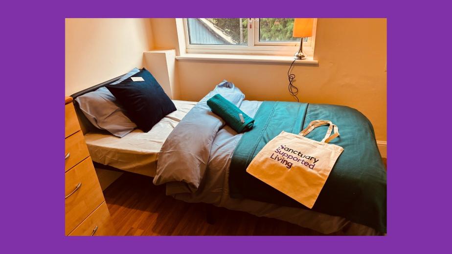 A cozy bedroom featuring a single bed with a blue and gray bedding set. A green towel is neatly placed on the bed, alongside a beige tote bag that reads "sanctuary supported living." A wooden dresser is visible on the left, and a window with natural light illuminates the room.