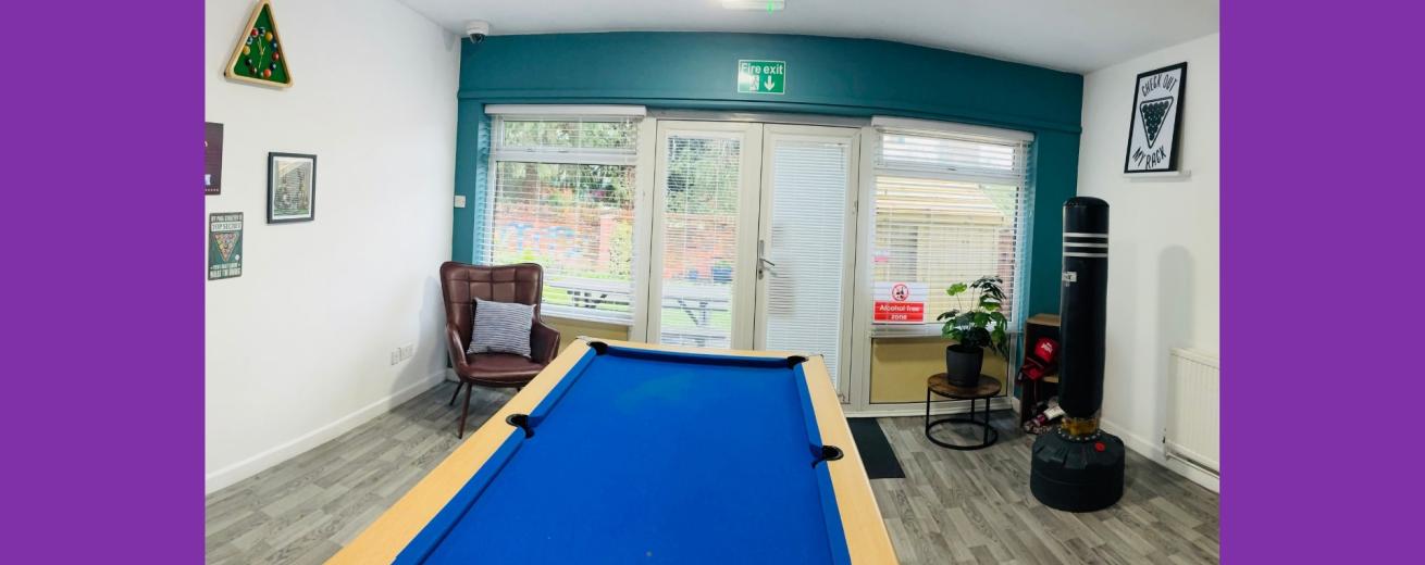 A spacious room featuring a blue pool table in the foreground. The walls are decorated with various framed posters and a chalkboard. A large window allows natural light to enter, revealing a view of an outdoor area. There is a comfortable armchair in one corner and a potted plant on a small table. A punching bag hangs from the ceiling, adding to the recreational atmosphere. The flooring is light-colored wood, contributing to a modern and inviting space.