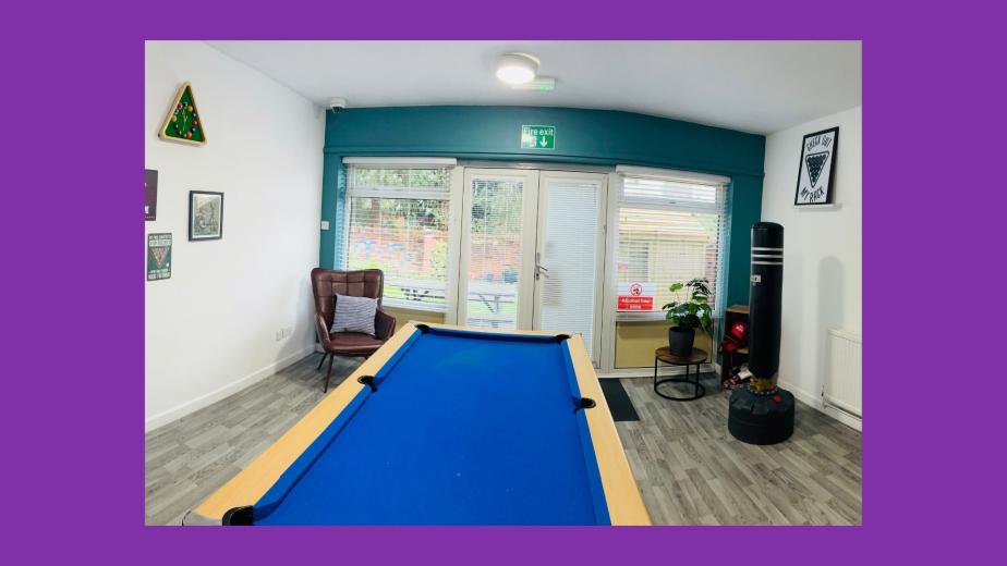 A spacious room featuring a blue pool table in the foreground. The walls are decorated with various framed posters and a chalkboard. A large window allows natural light to enter, revealing a view of an outdoor area. There is a comfortable armchair in one corner and a potted plant on a small table. A punching bag hangs from the ceiling, adding to the recreational atmosphere. The flooring is light-colored wood, contributing to a modern and inviting space.