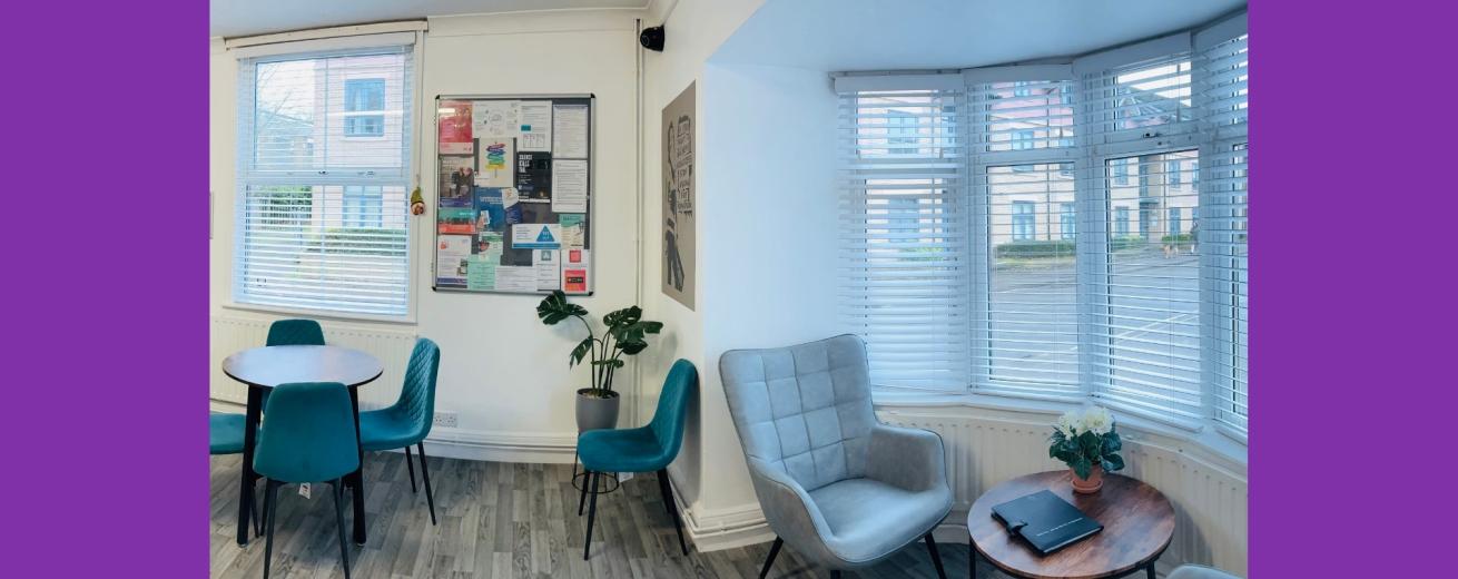 A spacious, well-lit communal area featuring two round wooden tables surrounded by teal chairs. On one side, there is a cozy gray armchair next to a small side table with a plant. The walls are adorned with various framed pictures and posters, and there are shelves with books and decorative items. Large windows allow natural light to fill the room, enhancing the inviting atmosphere.