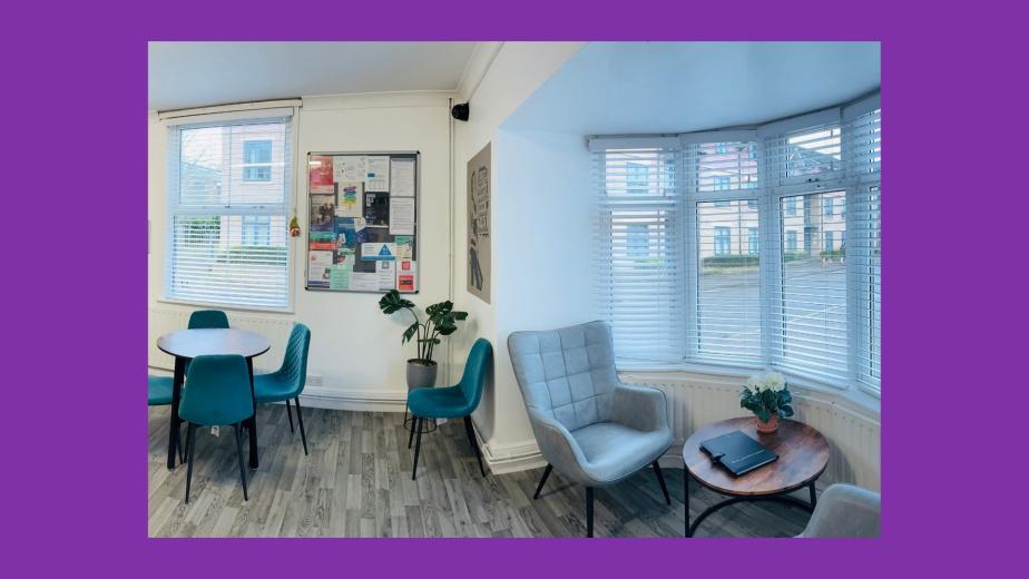 A spacious, well-lit communal area featuring two round wooden tables surrounded by teal chairs. On one side, there is a cozy gray armchair next to a small side table with a plant. The walls are adorned with various framed pictures and posters, and there are shelves with books and decorative items. Large windows allow natural light to fill the room, enhancing the inviting atmosphere.
