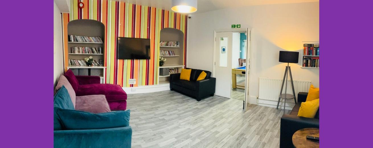 A cozy lounge area featuring a colorful striped accent wall. The room is furnished with two black sofas, one purple sofa, and a wooden coffee table. There are bookshelves on either side of a wall-mounted television, filled with books and decorative items. A floor lamp stands in one corner, and a door leads to another room. The flooring is light-colored wood, contributing to a warm and inviting atmosphere.