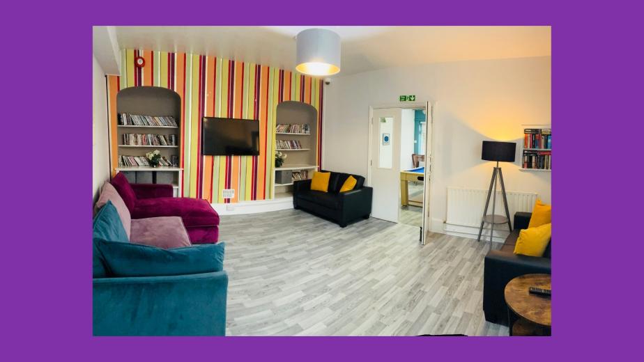 A cozy lounge area featuring a colorful striped accent wall. The room is furnished with two black sofas, one purple sofa, and a wooden coffee table. There are bookshelves on either side of a wall-mounted television, filled with books and decorative items. A floor lamp stands in one corner, and a door leads to another room. The flooring is light-colored wood, contributing to a warm and inviting atmosphere.