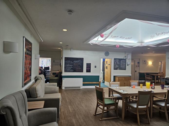 A spacious communal area featuring light-colored walls and wooden flooring. On the left, there are two comfortable armchairs facing a table set with colorful cups. In the center, a large table is surrounded by chairs, and a chalkboard displays handwritten notes. The ceiling has decorative elements, and a clock is visible on the wall. In the background, a doorway leads to another room where people can be seen.