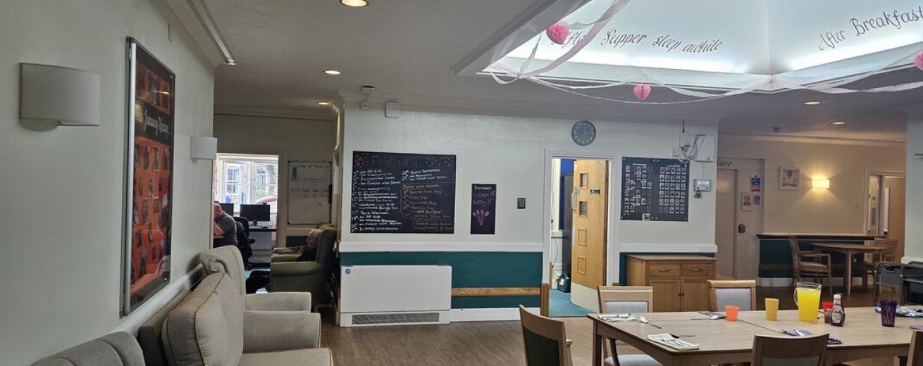 A spacious communal area featuring light-colored walls and wooden flooring. On the left, there are two comfortable armchairs facing a table set with colorful cups. In the center, a large table is surrounded by chairs, and a chalkboard displays handwritten notes. The ceiling has decorative elements, and a clock is visible on the wall. In the background, a doorway leads to another room where people can be seen.