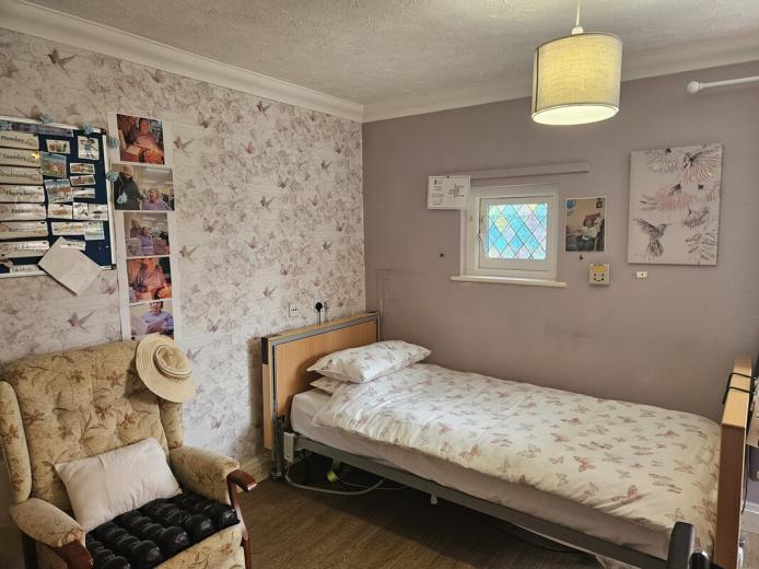 A cozy bedroom featuring a single bed with floral bedding against a light purple wall adorned with butterfly-patterned wallpaper. A small window with a colorful glass design allows natural light in. Next to the bed is a wooden bedside table. In the corner, there is a comfortable armchair with a patterned cushion and a sun hat resting on the arm. A bulletin board with various photos and notes is mounted on the wall, adding a personal touch to the space. 
