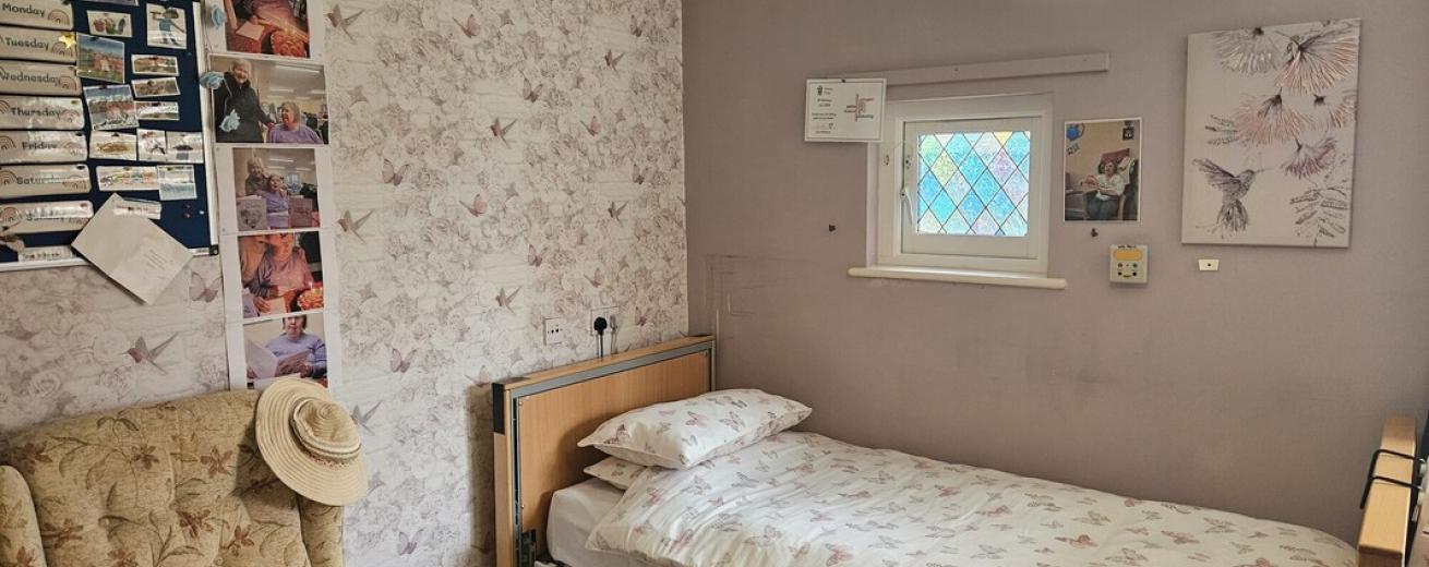 A cozy bedroom featuring a single bed with floral bedding against a light purple wall adorned with butterfly-patterned wallpaper. A small window with a colorful glass design allows natural light in. Next to the bed is a wooden bedside table. In the corner, there is a comfortable armchair with a patterned cushion and a sun hat resting on the arm. A bulletin board with various photos and notes is mounted on the wall, adding a personal touch to the space. 