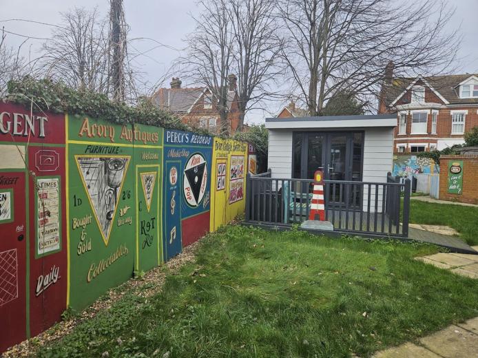 A colorful garden scene featuring a wooden shed with large windows and a small porch, surrounded by a grassy area. To the left, there is a vibrant mural on a fence showcasing various antique signs and advertisements, including references to furniture and collectibles. In the background, there are residential houses with trees and a cloudy sky. A traffic cone is placed near the porch, adding a quirky touch to the setting.