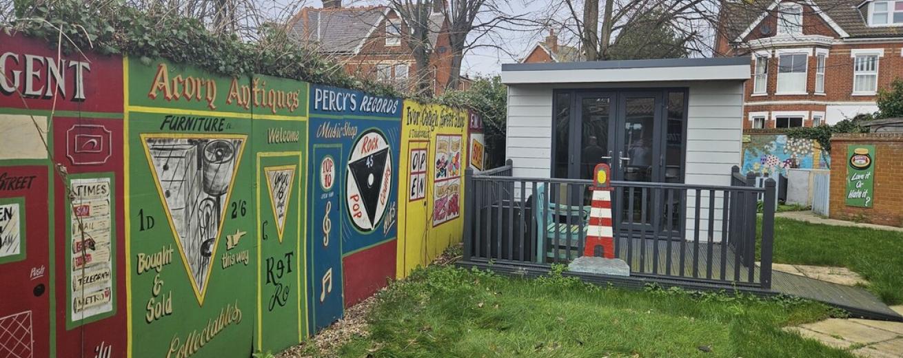 A colorful garden scene featuring a wooden shed with large windows and a small porch, surrounded by a grassy area. To the left, there is a vibrant mural on a fence showcasing various antique signs and advertisements, including references to furniture and collectibles. In the background, there are residential houses with trees and a cloudy sky. A traffic cone is placed near the porch, adding a quirky touch to the setting.