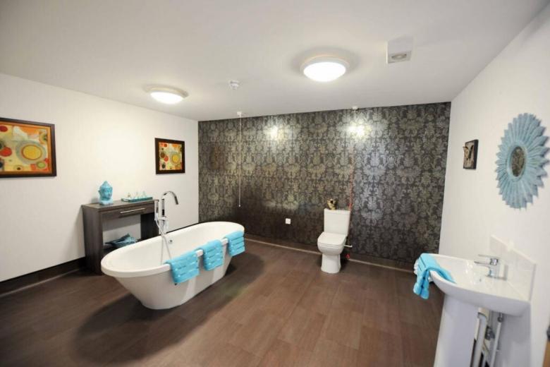 A modern bathroom features a freestanding bathtub with a blue towel and a small table with a statue and a framed print. A white toilet sits on a dark brown tiled floor. The walls are covered in a dark, patterned wallpaper. Two round mirrors with a blue frame are mounted on the wall.