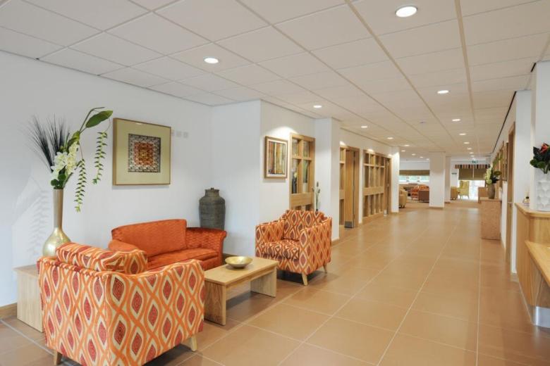 Interior view of a waiting area with two patterned orange armchairs and a small table, flanked by walls with artwork and a hallway leading into a brighter room.
